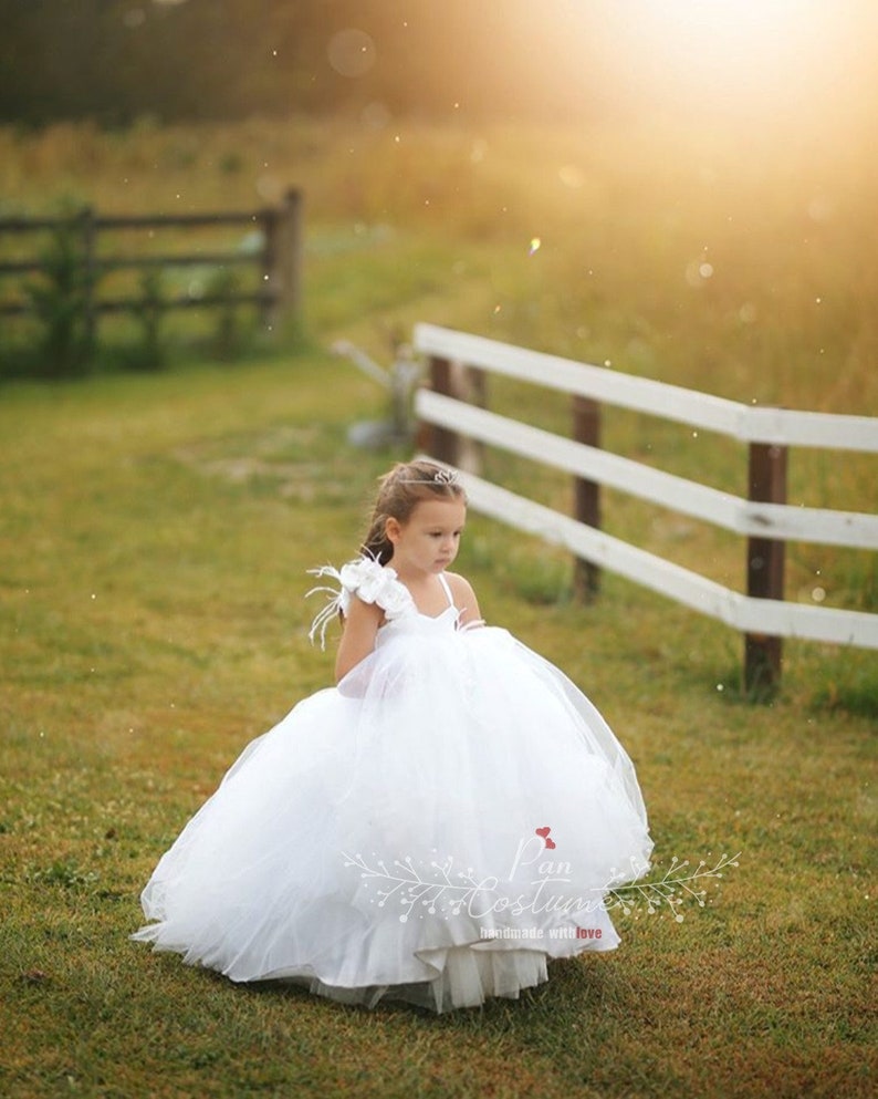 May include: A young girl wearing a white tulle dress with a single white flower and feather on the shoulder. The dress has a sweetheart neckline and a full skirt. The girl is standing in a grassy field with a white fence in the background.