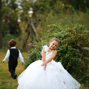 May include: A young girl in a white tulle dress with a large white flower on the shoulder. She is smiling and looking at the camera. The dress has a white sash and a white flower on the front. The girl is standing in a grassy field with a boy in the background.