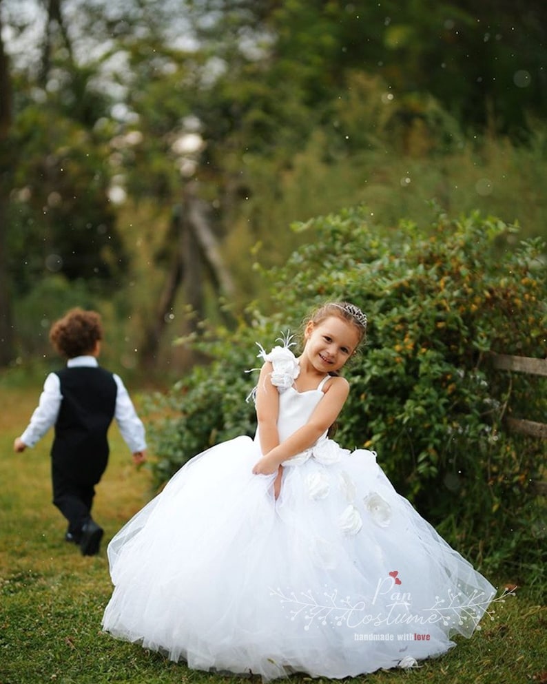May include: A young girl in a white tulle dress with a large white flower on the shoulder. She is smiling and looking at the camera. The dress has a white sash and a white flower on the front. The girl is standing in a grassy field with a boy in the background.