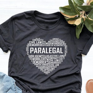 May include: Black t-shirt featuring a white graphic design of the word "Paralegal" surrounded by legal terms. The shirt is laid flat on a white surface with a pair of brown sandals.