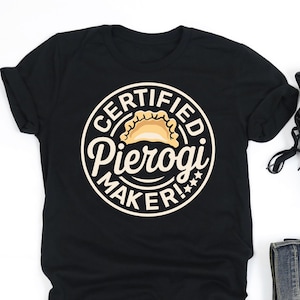 May include: Black t-shirt with a circular design that reads "CERTIFIED Pierogi MAKER!" in a white font. The design includes an illustration of a pierogi. The shirt is paired with black sneakers and blue jeans.