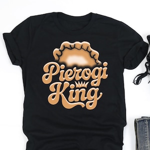 May include: Black t-shirt with the words "Pierogi King" in a retro font and a pierogi graphic. The image also includes black sneakers, folded denim jeans, and wireless earbuds.