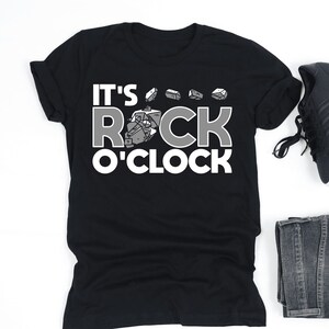 May include: Black t-shirt with the phrase "IT'S ROCK O'CLOCK" in white and gray lettering, accompanied by rock illustrations. The image also includes black sneakers, folded denim jeans, and wireless earbuds.