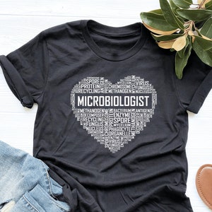 May include: Black t-shirt with a white heart-shaped design featuring the word "MICROBIOLOGIST" surrounded by terms related to microbiology, such as "bacteria", "virus", "fungus", and "spore".