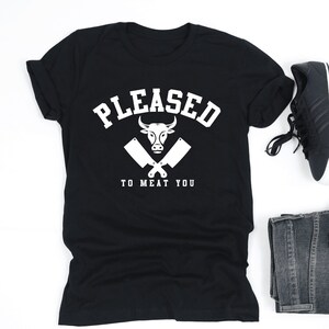 May include: Black t-shirt with a white graphic of a bull's head with crossed butcher knives and the text "PLEASED TO MEAT YOU".