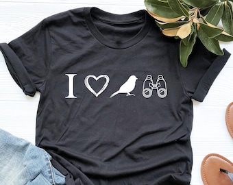 May include: Black t-shirt with white text that reads "I love birds" with a heart and a bird graphic and binoculars. The shirt is laying on a white surface with a pair of blue jeans and a green plant.