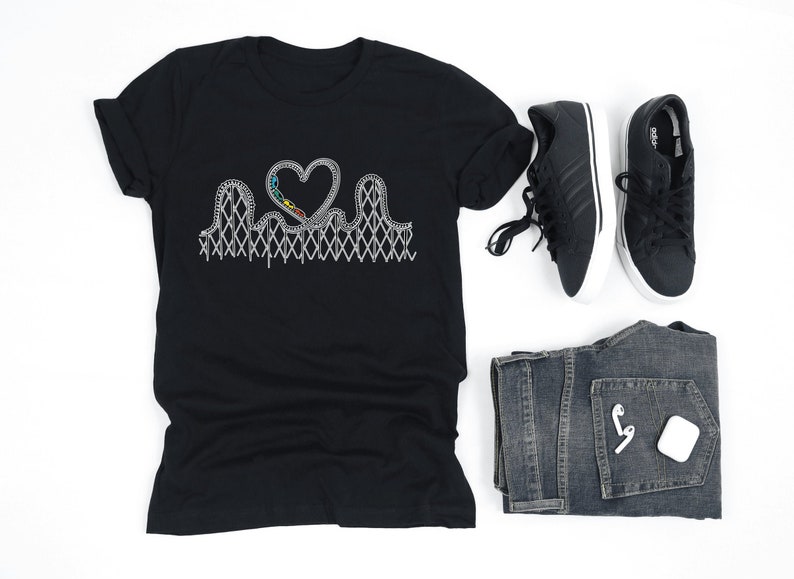 May include: Black t-shirt featuring a roller coaster design in white, forming a heart shape. The shirt is paired with black sneakers, blue jeans, and white wireless earbuds. A casual, everyday outfit.