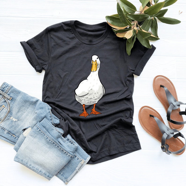 Puede incluir: Una camiseta negra con un gr&aacute;fico de un pato blanco. El pato est&aacute; mirando hacia el lado derecho de la imagen y tiene un pico y patas naranjas.