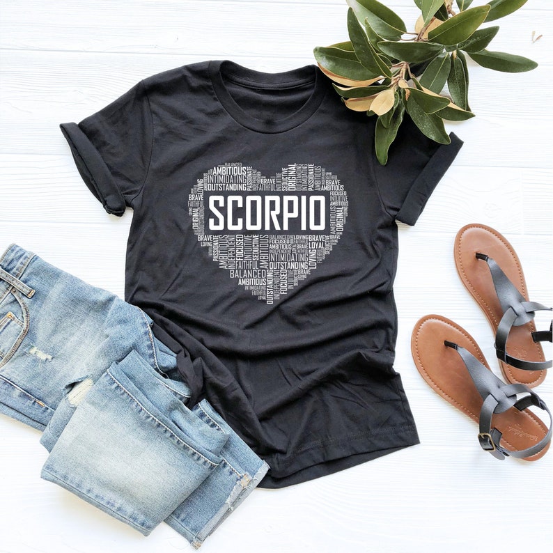 May include: Black t-shirt with a white heart-shaped word cloud design featuring the word "SCORPIO" in the center. The word cloud includes words like "ambitious", "focused", "original", "brave", "loyal", and "intimidating".
