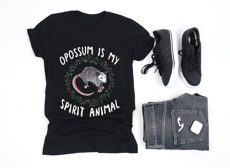 May include: Black t-shirt with white text that reads "Opossum is my spirit animal". A cartoon opossum is illustrated inside a wreath. The t-shirt is laying flat on a white surface.