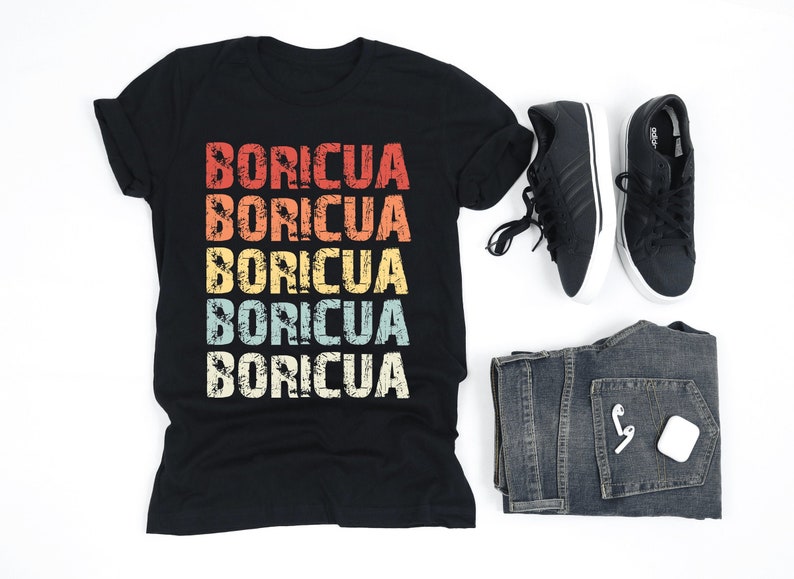 Może przedstawiać: Czarny T-shirt z napisem "BORICUA" powt&oacute;rzonym pięć razy w vintage'owym schemacie kolor&oacute;w tęczy. Koszulka leży płasko na białym tle z parą czarnych but&oacute;w sportowych i parą niebieskich dżins&oacute;w.