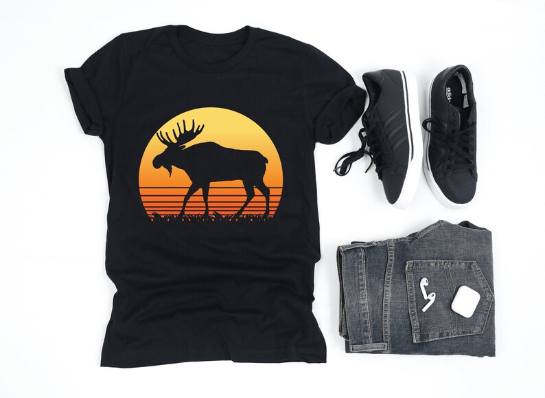 May include: Black t-shirt featuring a silhouette of a moose against a sunset design. The image also includes black sneakers, blue jeans, and wireless earbuds. A casual outfit with a nature theme.