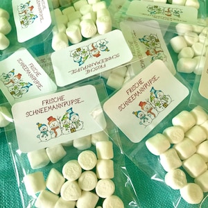 Könnte beinhalten: Mehrere kleine Tüten mit weißen Marshmallows mit einem bedruckten Etikett, auf dem "Frische Schneemannpupse..." steht, was im Deutschen "Frische Schneemannpupse..." bedeutet.