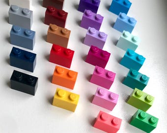 May include: A collection of twenty-eight rectangular plastic building bricks arranged in four vertical columns. Each brick features two circular studs on the top surface. The bricks are presented in a wide spectrum of colors including white, gray, blue, black, brown, red, orange, yellow, purple, pink, and various shades of green and teal.