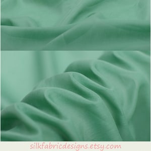 Könnte beinhalten: Eine Nahaufnahme eines hellgrünen Stoffes mit einer glatten, seidenartigen Textur. Der Stoff ist drapiert und gefaltet, um seinen Fall und seine Fließfähigkeit zu zeigen. Der Stoff ist auf silkfabricdesigns.etsy.com erhältlich.