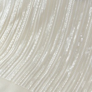 Può includere: Tessuto bianco trasparente con file verticali di paillettes. Le paillettes sono piccole e distanziate uniformemente.