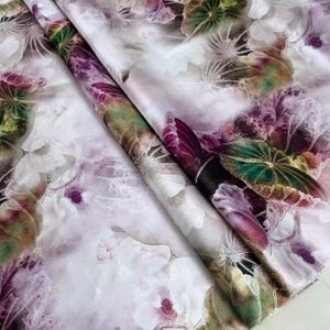 May include: A close-up of a folded fabric with a floral pattern. The design features white, purple, and green flowers and leaves on a light background. The fabric has a silky sheen and a soft, flowing drape.