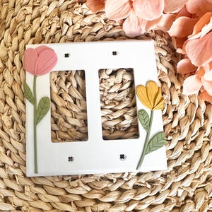 May include: White double light switch cover decorated with a pink and yellow flower design. The flowers have green stems and leaves. The cover is set against a woven background with pink flowers.