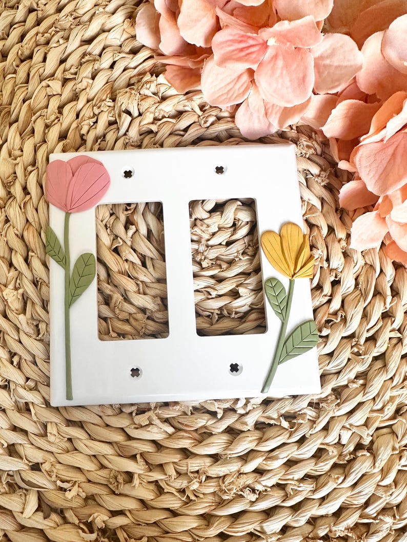 May include: White double light switch cover decorated with a pink and yellow flower design. The flowers have green stems and leaves. The cover is set against a woven background with pink flowers.
