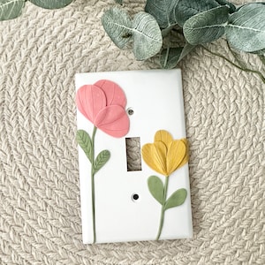 以下が含まれることがあります： ピンクと黄色の花柄の白いライトスイッチカバー。花は粘土でできており、緑色の茎と葉があります。