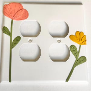 May include: White electrical outlet cover with two decorative flowers. One flower is orange, the other is yellow. Both flowers have green stems and leaves. The cover has four outlet holes.
