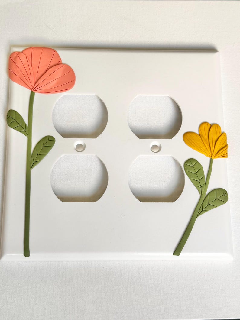 May include: White electrical outlet cover with two decorative flowers. One flower is orange, the other is yellow. Both flowers have green stems and leaves. The cover has four outlet holes.