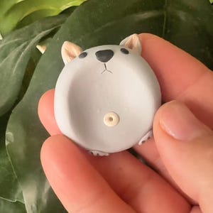 May include: A grey ceramic koala-shaped trinket dish. The dish has a small, round body with a recessed area for holding small items. The koala has small, tan ears, a black nose, and a small, off-white circle on its belly.