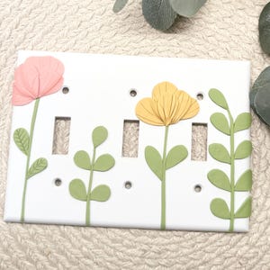 May include: White light switch cover decorated with pink and yellow flowers and green leaves. The floral design is 3D and adds a decorative touch to any room. The cover has three switch openings and small screw holes.