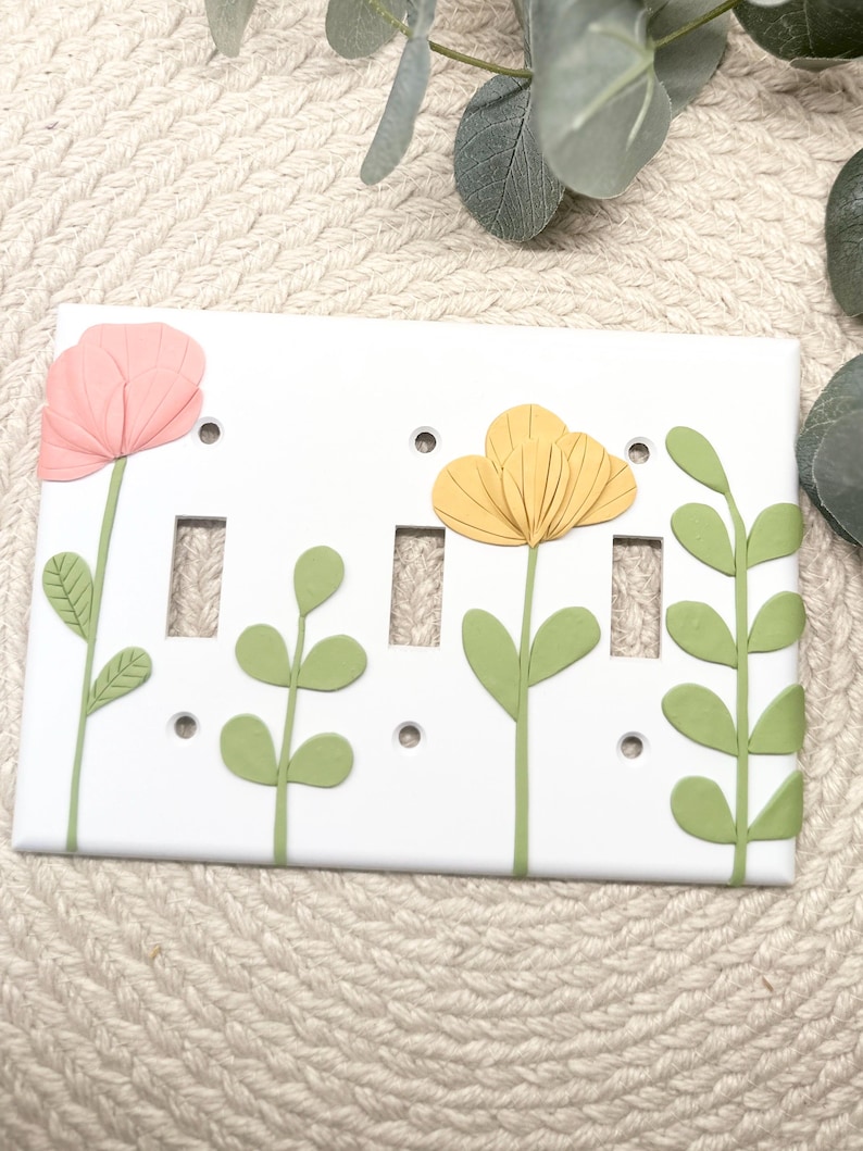 May include: White light switch cover decorated with pink and yellow flowers and green leaves. The floral design is 3D and adds a decorative touch to any room. The cover has three switch openings and small screw holes.