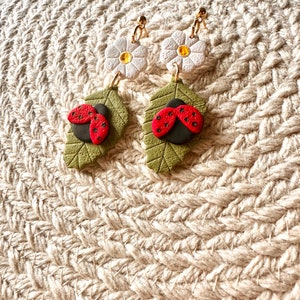Puede incluir: Un par de pendientes con una mariquita en una hoja verde y una flor blanca con un centro amarillo.