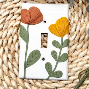 Pode incluir: Uma tampa de interruptor de luz branca com um desenho pintado à mão de duas flores estilizadas. As flores são laranja e amarelas com folhas verdes.