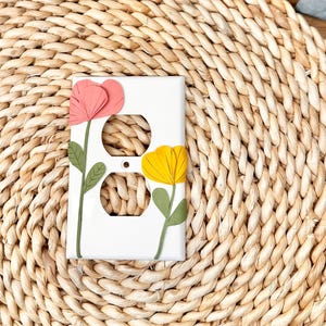 May include: White electrical outlet cover decorated with a pink and yellow flower design. The flowers have green stems and leaves. The cover is rectangular and has two outlet holes.