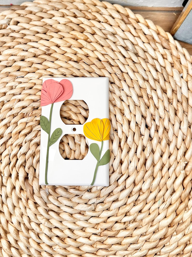 May include: White electrical outlet cover decorated with a pink and yellow flower design. The flowers have green stems and leaves. The cover is rectangular and has two outlet holes.