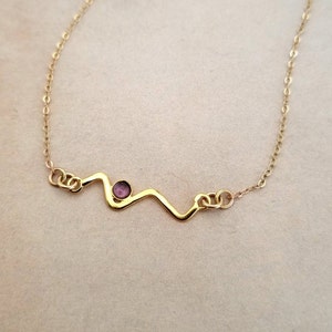 May include: Delicate gold-tone necklace featuring a wavy design with a small, round, purple gemstone at its center. The necklace has a fine chain and is displayed on a neutral background. A minimalist piece of jewelry.