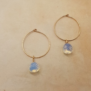 Op de afbeelding: Een paar gouden creolen met een delicate, traanvormige opaalhanger die aan elke creool hangt.