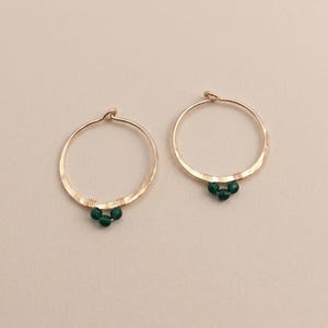 Op de afbeelding: Een paar goudkleurige oorringen met een gehamerde metaaltextuur. Elke ring is aan de onderkant versierd met drie kleine, ronde, donkergroene kralen die zijn vastgezet met dun goudkleurig draad voor een verfijnd en gebalanceerd ontwerp.