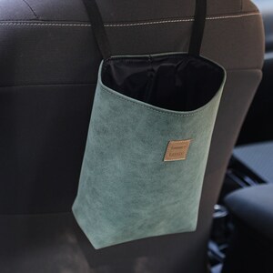 May include: A teal car tidy bag suspended from a car seat headrest. The bag is made of a soft, suede-like material with a black interior. A small, rectangular brown label with text is attached to the front.