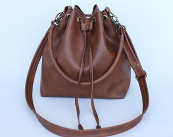 May include: A brown leather bucket bag with a drawstring closure and adjustable shoulder strap. The bag has a textured surface and metal hardware. The strap is attached with metal clasps and rivets. The bag is designed to be carried over the shoulder or by the handles.