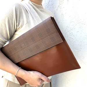 May include: A brown leather clutch with a plaid flap. The clutch is held by a person. The leather is a rich, warm brown, and the flap features a classic brown and black plaid pattern. The clutch appears to be a large size, suitable for carrying documents or a tablet.