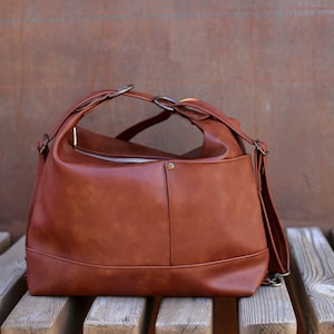 Può includere: Una borsa a tracolla in pelle marrone con manico superiore e tracolla staccabile. La borsa ha una chiusura superiore con cerniera e una tasca frontale. La borsa è appoggiata su una panchina di legno.