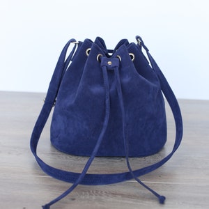 May include: A navy blue suede bucket bag with a drawstring closure and a long shoulder strap. The bag has gold-coloured metal accents and a soft, textured appearance. The bag is sitting on a wooden surface.