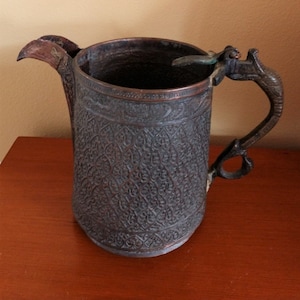 May include: A copper pitcher with an ornate design and a hinged lid. The pitcher has a handle shaped like a bird's head.