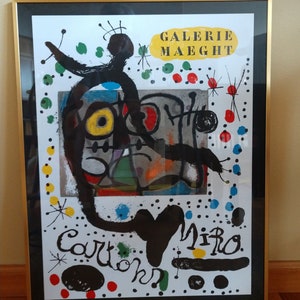 May include: Framed Miro lithograph poster. Black, white, red, yellow, blue, and green colors.  The poster features abstract figures and text: 'Galerie Maeght', 'Miro', and 'Cartoon'. The artwork is bold and expressive, characteristic of Miro's style.