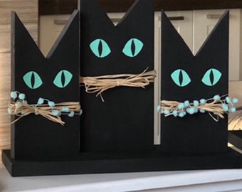 May include: A black wooden cat decoration with three cat silhouettes. Each cat has light blue eyes and is adorned with twine or a string of blue beads. The cats are mounted on a black base.