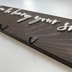 May include: A wooden sign with the text "Place to hang your shoes" in white raised letters. The sign has three black metal hooks.