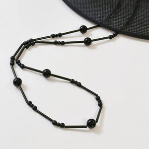 May include: Black beaded necklace with a matching black hat. The necklace features a series of small and large black beads strung on thin black rods. The hat has a mesh design.
