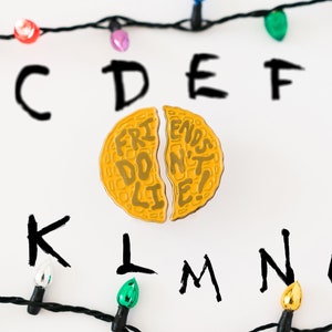 May include: A yellow enamel pin shaped like a waffle with the text "Friends Don't Lie!" on it. The pin is split in half, with the text "Friends Don't" on one half and "Lie!" on the other. The pin is surrounded by black string lights with colored bulbs and the letters of the alphabet, B through O, written in black marker on a white background.