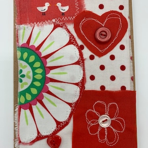 Könnte beinhalten: Handgemachte Grußkarte mit Stoffflicken. Verfügt über ein rotes Herz mit einem Knopf, ein Blumendesign und ein Tupfenmuster. Enthält ein Blumendesign und zwei Vögel. Die Karte besteht aus einer braunen Kartonbasis.