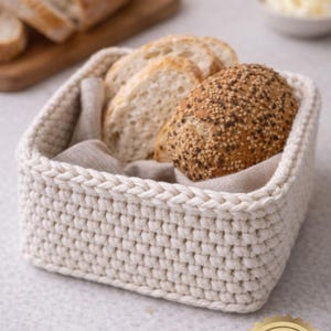 May include: A square, off-white crochet basket filled with sliced bread and a sesame seed roll. The basket sits on a light surface, with a wooden cutting board and a small bowl of butter in the background.