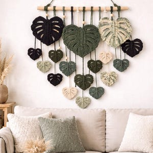May include: A macrame wall hanging featuring a series of leaf-shaped designs in various shades of green, black, and cream. The leaves are suspended from a wooden dowel with black cords. The text "PDF-BEGINNER-INTERMEDIATE" is at the top.